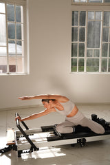 Žena u Être Active Fit light gray sports bra i high-waisted leggings - pilates reformer stretching vježba, bočni pogled, indoor studio s velikim prozorima i dramatičnim prirodnim svjetlom, premium sportska odjeća za pilates