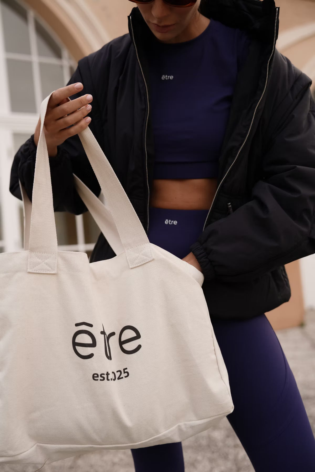 Navy plavi Contour Fit sportski set Être - crop top i tajice s logom, styling s bež tote bag torbom est.025 i crnom bomber jaknom, outdoor lifestyle fotografija, premium ženska sportska odjeća