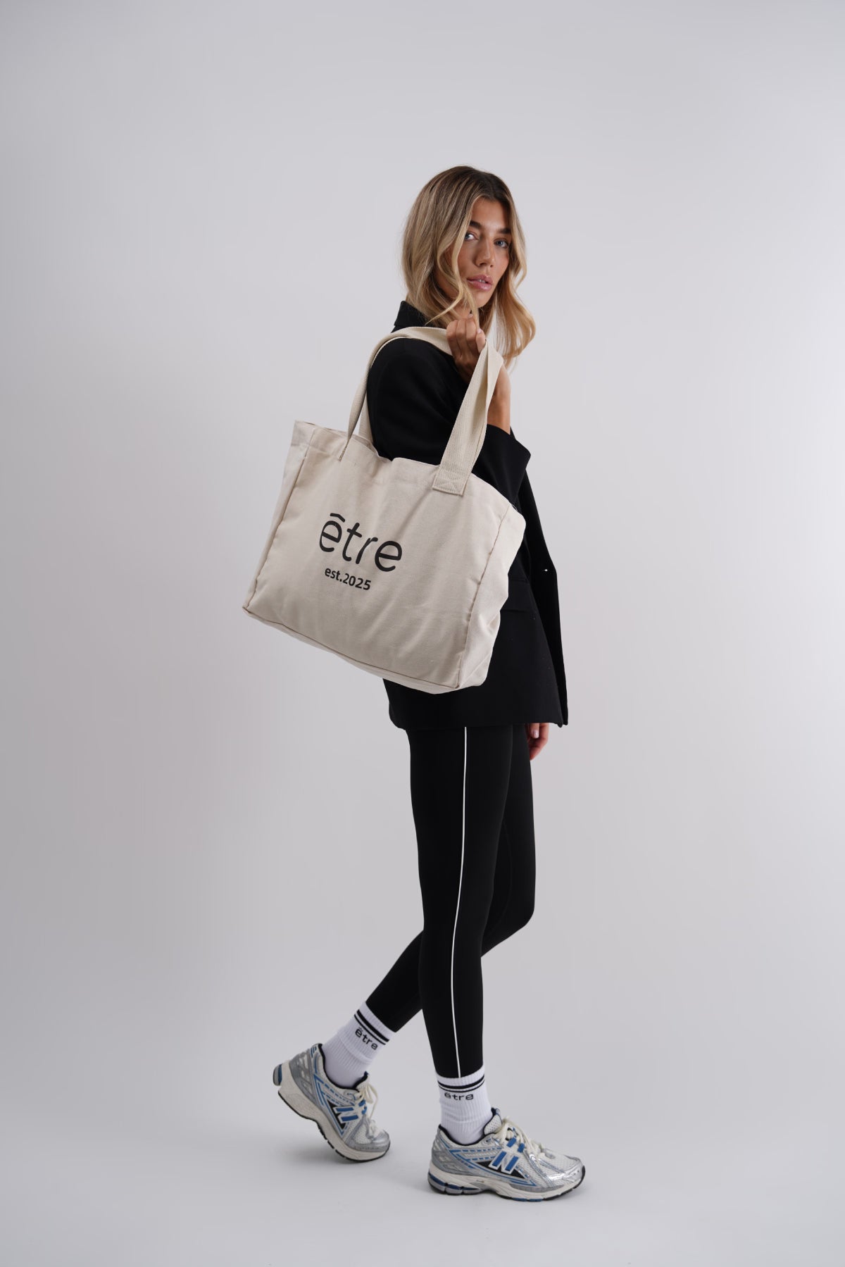 Bež Être tote bag platnena torba - lifestyle fotografija s kompletnim Être outfitom, crne tajice i bijele sportske čarape, shopping bag s logom est.2025